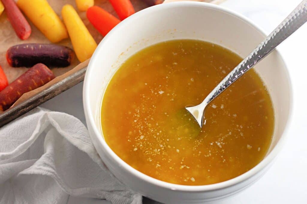 A white bowl filled with golden-yellow sauce or dressing with a spoon inside, placed next to a tray of colorful carrots and a white cloth.