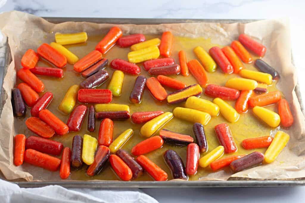 A baking sheet lined with parchment paper holds a mix of colorful roasted baby carrots in orange, yellow, and purple hues, coated in oil.