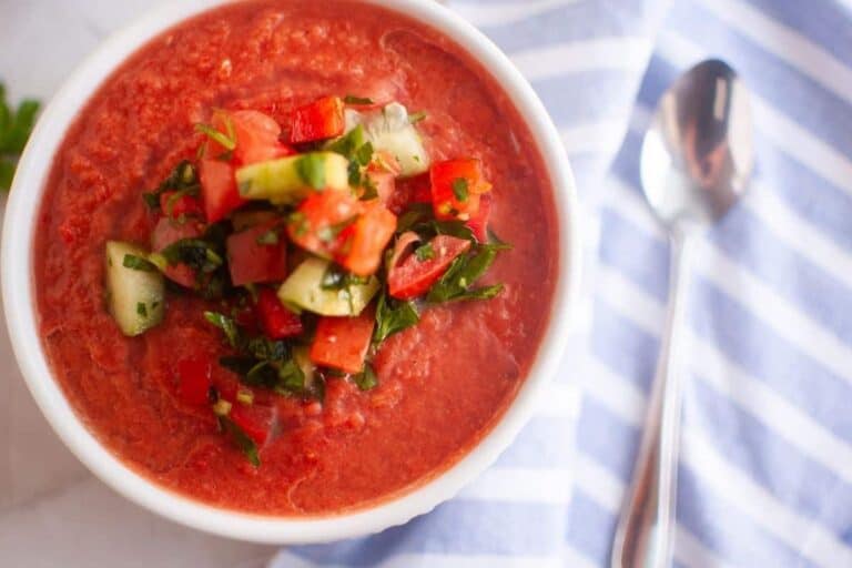 A bowl of gazpacho topped with chopped cucumber, tomato, and herbs, next to a silver spoon on a blue and white striped cloth.