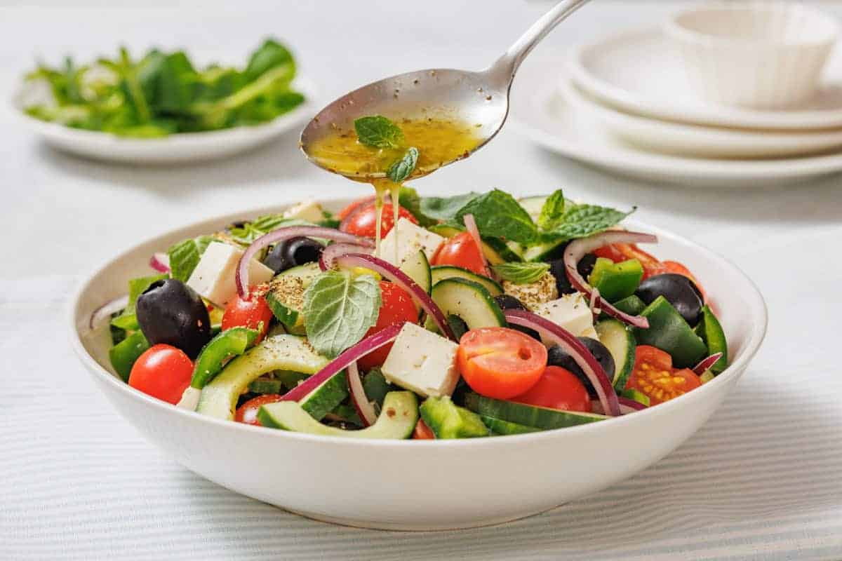 A spoon drizzles dressing over a bowl of Farmhouse Veggie Salad with Feta.