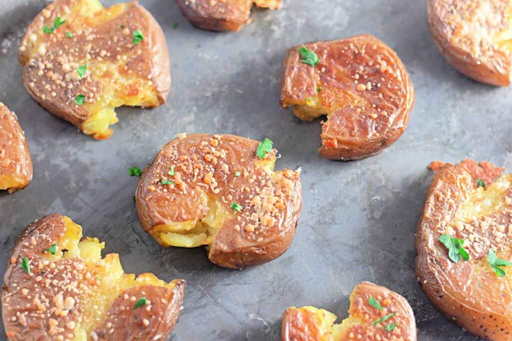 Crispy Garlic Parmesan Smashed Potatoes topped with herbs and grated cheese on a metal baking sheet.
