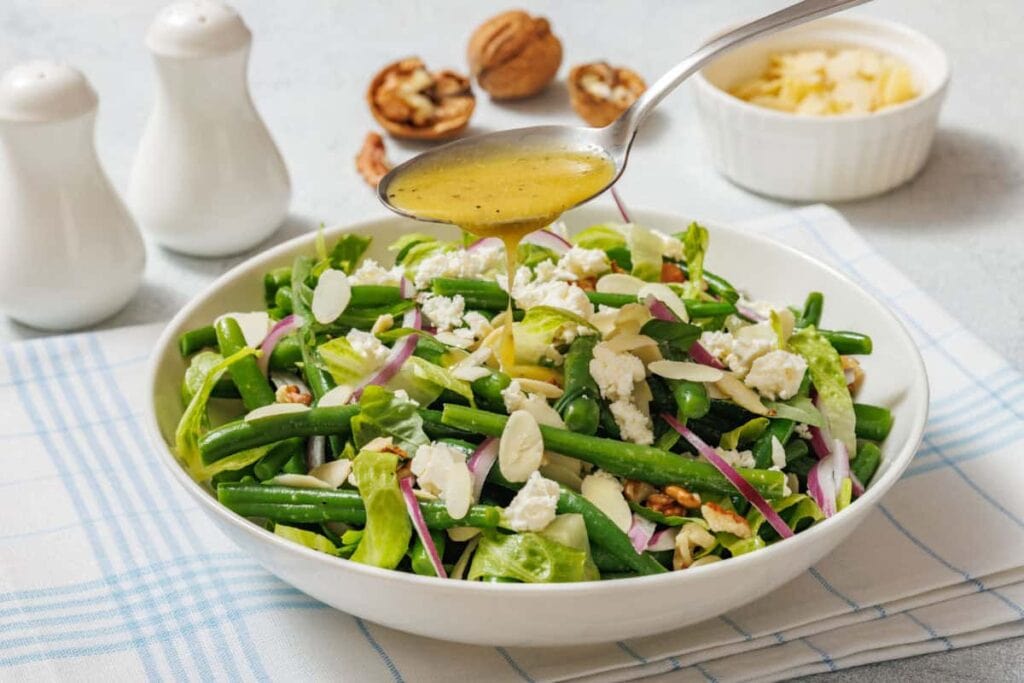 A bowl of green bean salad with feta, red onion, and sliced almonds, with dressing being poured from a spoon. Salt and pepper shakers and walnuts are in the background.