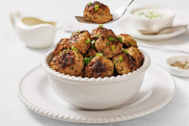 A white bowl filled with grilled meatballs, garnished with chopped green herbs, sits on a white plate; a fork holds one meatball above the bowl.