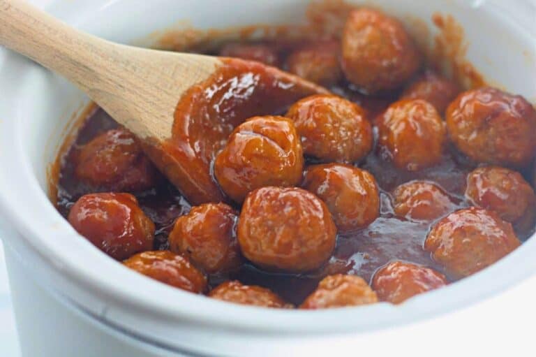 A wooden spoon stirs meatballs coated in thick red sauce inside a white slow cooker.