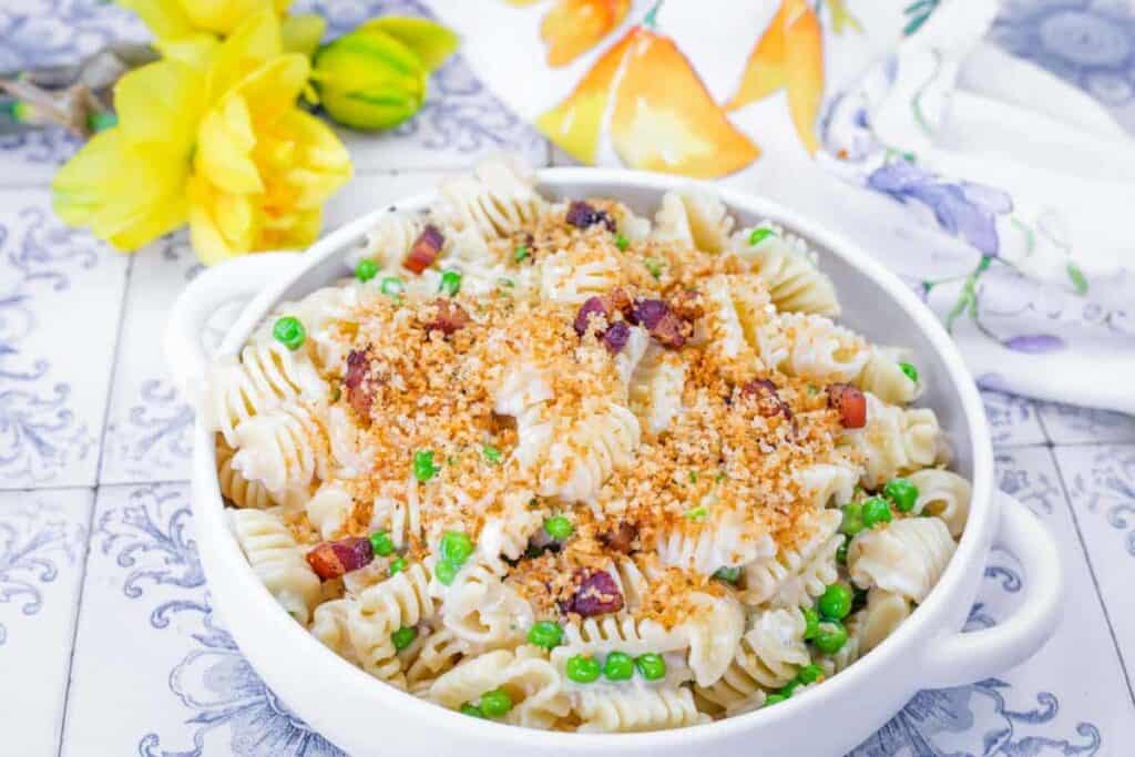 A bowl of spiral pasta with peas, bacon bits, and breadcrumbs sits on a tiled surface, with yellow flowers and a patterned napkin in the background.