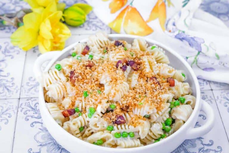 A bowl of spiral pasta with peas, bacon bits, and breadcrumbs sits on a tiled surface, with yellow flowers and a patterned napkin in the background.