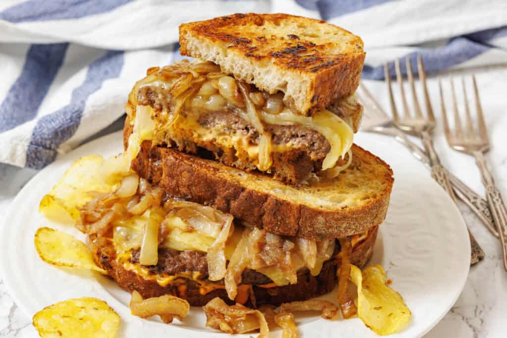 A patty melt sandwich with grilled onions and melted cheese on toasted bread, stacked on a plate with potato chips and forks in the background.