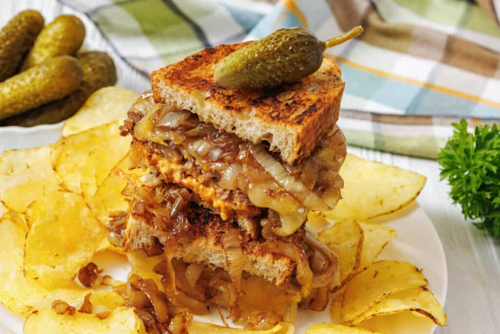 Stacked sandwich with caramelized onions and melted cheese on toasted bread, topped with a pickle, served with potato chips on a white plate.