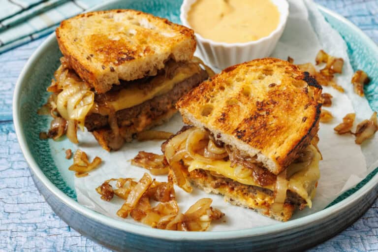 A plate with a classic patty melt cut in half, served with a cup of dipping sauce.