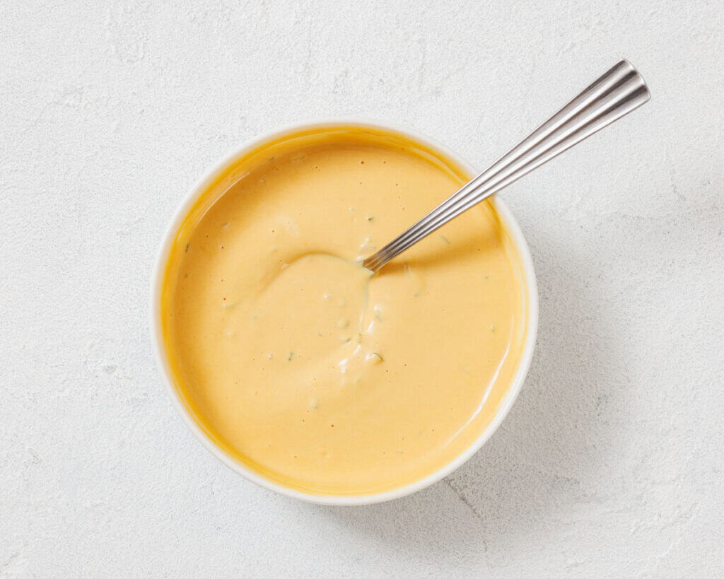 A white bowl filled with creamy yellow cheese sauce, with a metal spoon resting inside, on a light textured surface.