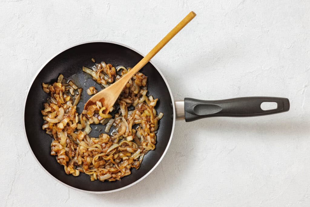 A frying pan with saut&eacute;ed, caramelized onions and a wooden spoon on a light surface.