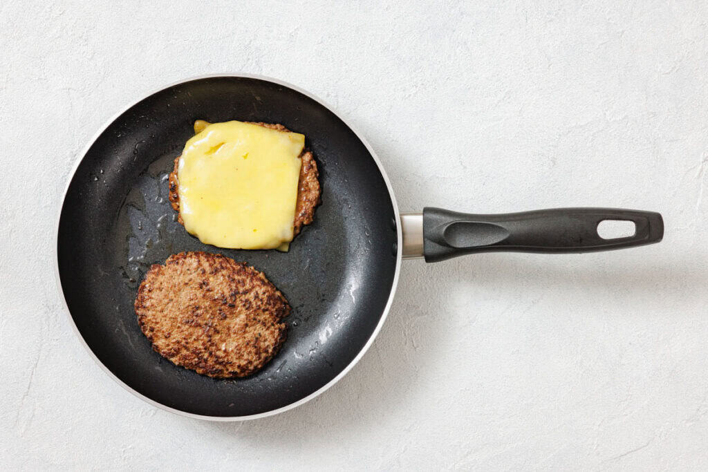 Two beef burger patties are cooking in a frying pan; one patty is topped with melted cheese, and the other is plain.