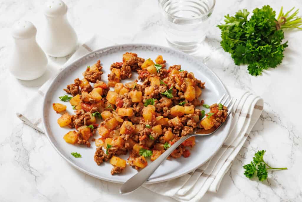 A slightly high-angle shot of a white plate holding a serving of "Poor Man's Meal," a savory mixture of browned ground meat, diced potatoes, and chopped tomatoes, garnished with fresh chopped parsley, with a silver fork resting on the plate.