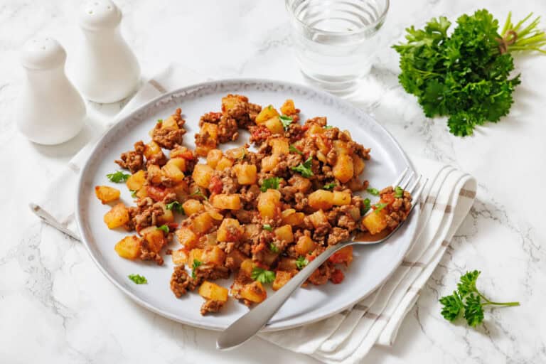A slightly high-angle shot of a white plate holding a serving of poor man's meal, a savory mixture of browned ground meat, diced potatoes, and chopped tomatoes, garnished with fresh chopped parsley.