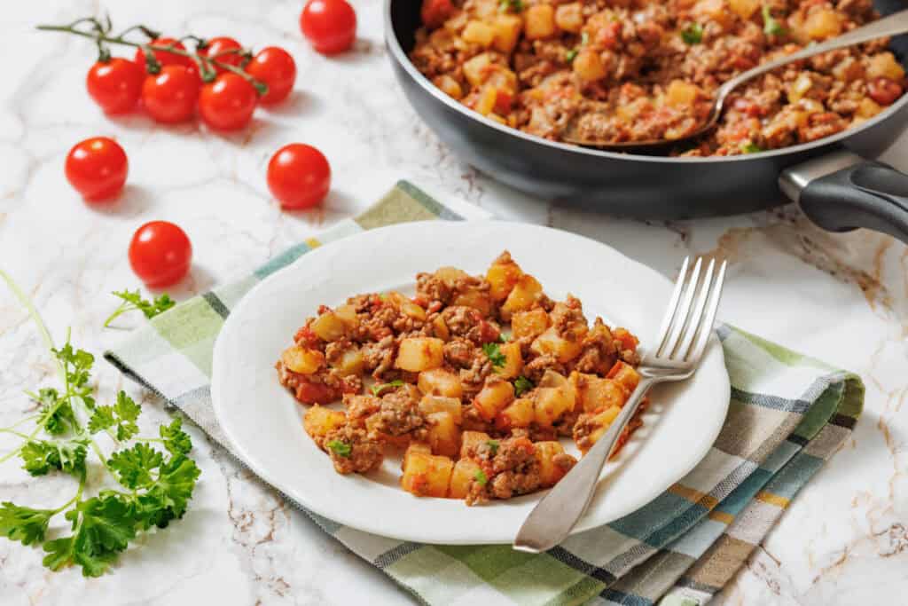 A close-up of a white plate holding a serving of "Poor Man's Meal," a savory mixture of browned ground meat, diced potatoes, and chopped tomatoes, garnished with fresh parsley, with a silver fork resting on the plate.