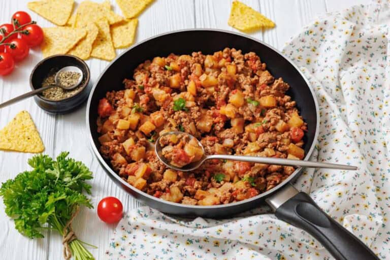 A frying pan filled with Poor Man's meal sits on a white surface with a spoon, surrounded by fresh parsley, tomatoes, tortilla chips, and seasoning.