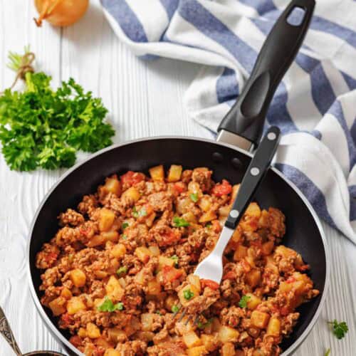 An overhead, slightly angled shot of a black non-stick skillet filled with a savory mixture of cooked ground meat, diced potatoes, and chopped tomatoes.