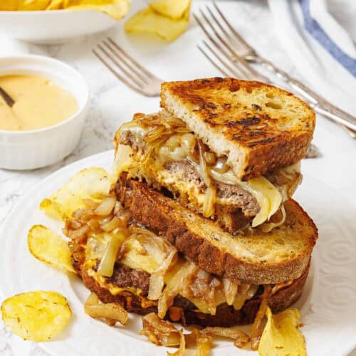 A plate with a stacked grilled cheese and caramelized onion sandwich, surrounded by potato chips, with a bowl of sauce and more chips in the background.
