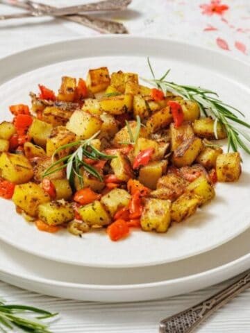A white plate with Skillet Fried Potatoes garnished with rosemary sits on a white table with cutlery and a small bowl of pepper on the side.
