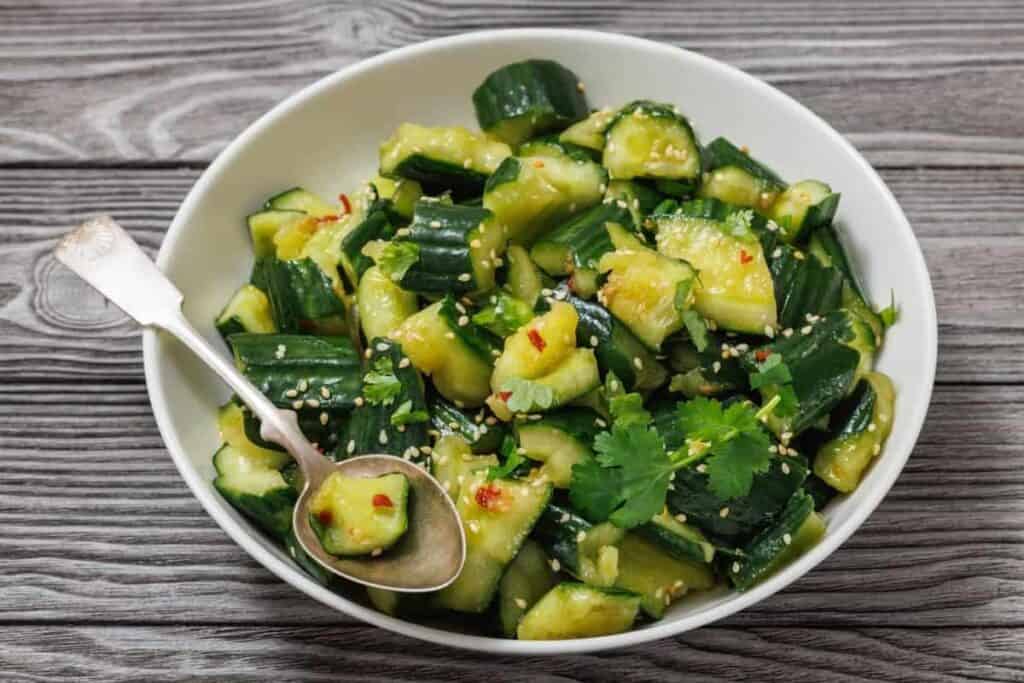 A white bowl filled with smashed cucumber salad garnished with cilantro, sesame seeds, and chili flakes, with a metal spoon resting inside. The bowl is on a wooden surface.