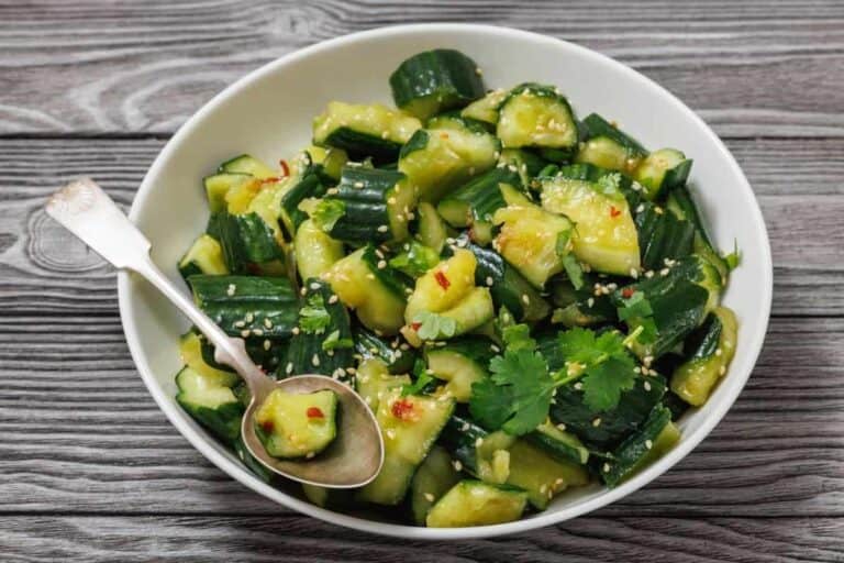 A white bowl filled with smashed cucumber salad garnished with cilantro, sesame seeds, and chili flakes, with a metal spoon resting inside. The bowl is on a wooden surface.