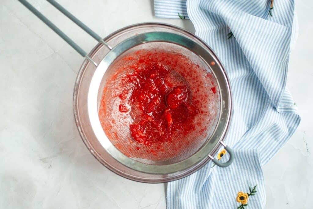 An overhead shot of a fine-mesh metal sieve, resting over a clear glass bowl, containing a thick, chunky red strawberry puree with some liquid already collected in the bowl below, with a blue and white striped napkin to the right.
