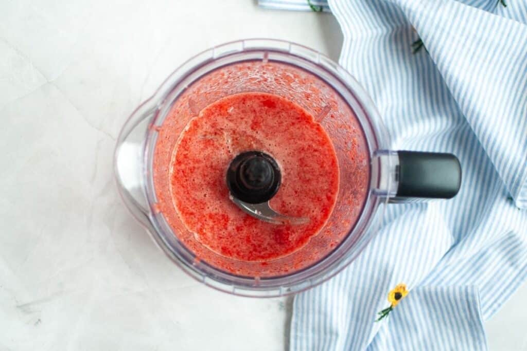 An overhead view of a food processor bowl containing a vibrant red, partially pureed mixture of strawberries, with a blue and white striped napkin to the right.