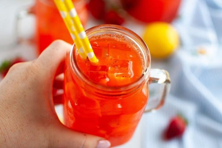 A close-up of a hand holding a clear glass with a handle, filled with bright orange-red strawberry lemonade and ice cubes, with two yellow and white striped paper straws.