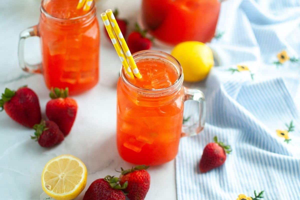 Glass mason jars filled with iced strawberry lemonade on a table, surrounded by fresh strawberries, a halved lemon, and a blue floral cloth.