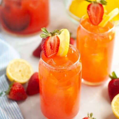 A close-up of two clear glasses of bright orange-red strawberry lemonade with ice, one in the foreground garnished with a halved strawberry and a lemon slice, with a pitcher of the drink, and scattered fresh strawberries and lemons in the blurred background.