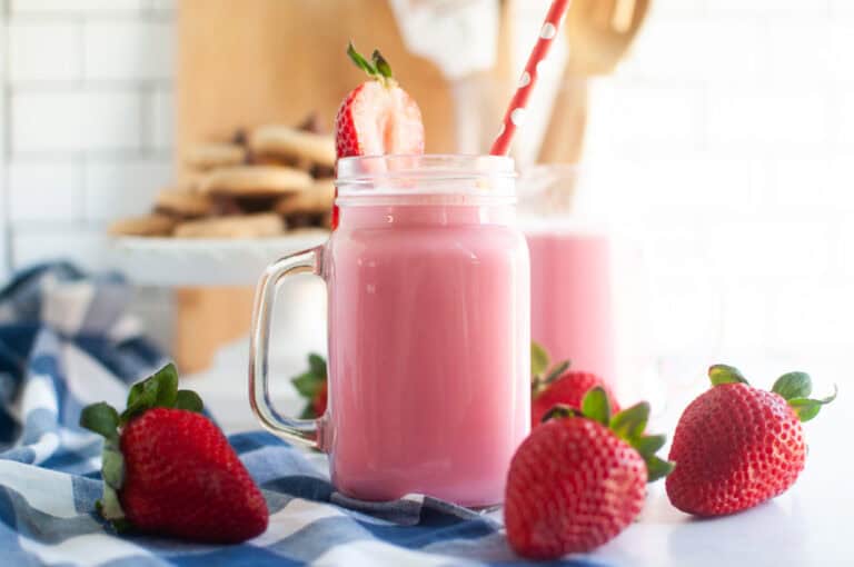 A glass jar filled with pink strawberry milk, garnished with a strawberry and a red straw, surrounded by fresh strawberries on a blue checkered cloth.