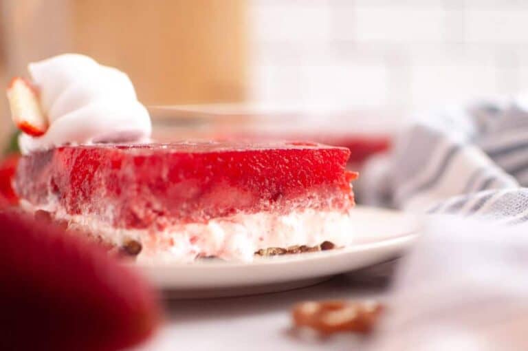 A slice of layered strawberry dessert with a red gelatin top and creamy middle, garnished with whipped cream and a strawberry, served on a white plate.