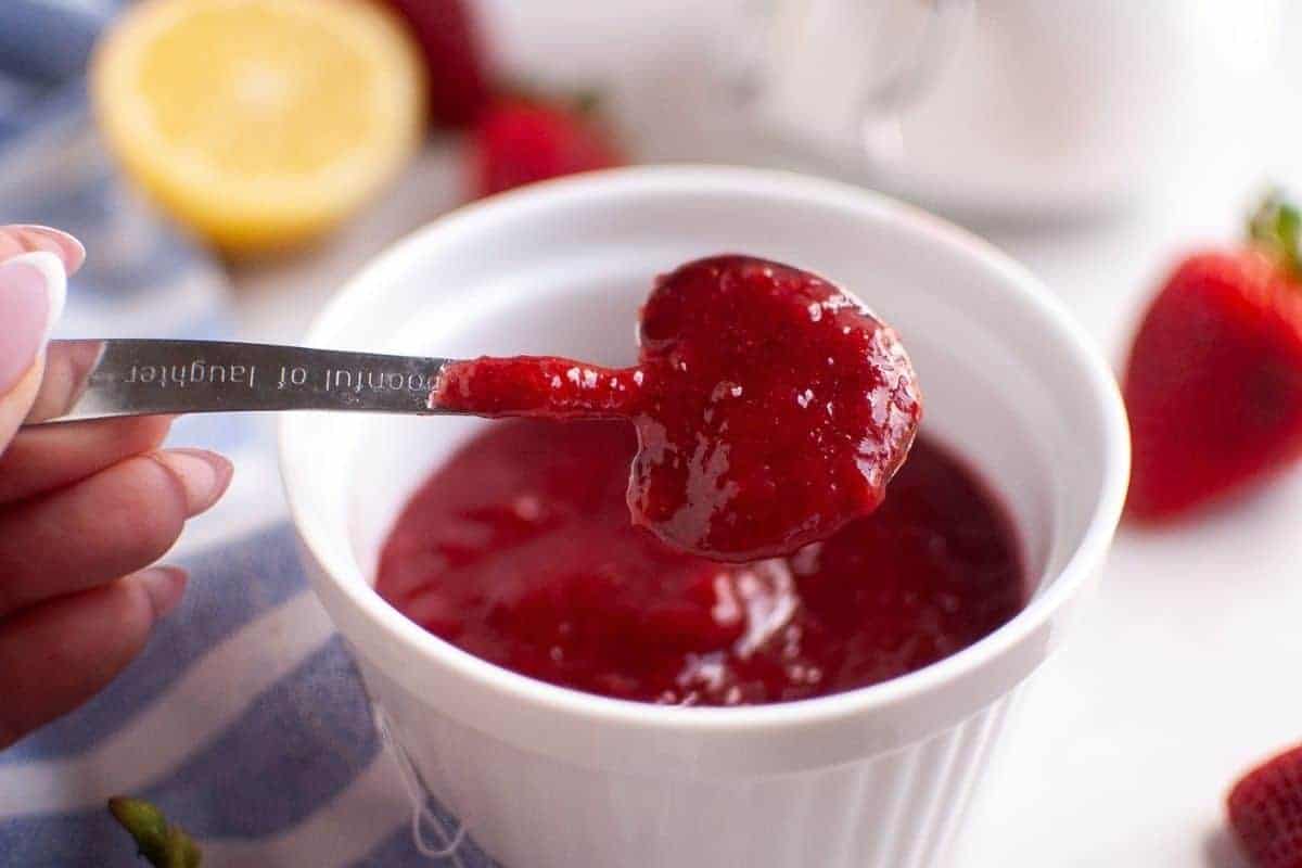 A hand holds a heart-shaped spoon filled with vibrant red strawberry puree above a white ramekin containing more puree, with fresh fruit and a lemon in the background.