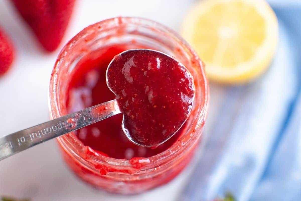 A heart-shaped measuring spoon holds a scoop of vibrant red strawberry puree over a glass jar filled with more puree, surrounded by fresh strawberries and a lemon half.