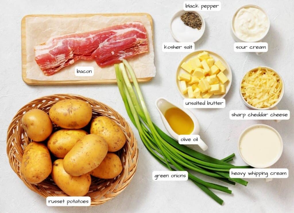Ingredients for loaded mashed potatoes are arranged on a white surface, including bacon, russet potatoes, green onions, olive oil, heavy cream, cheddar cheese, butter, sour cream, salt, and pepper.