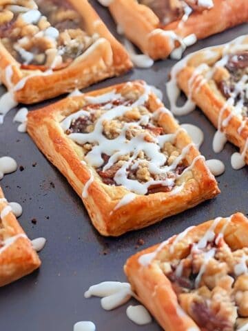 Golden-brown Dutch apple danishes topped with fruit, crumble topping and drizzled with white frosting on a dark baking sheet.