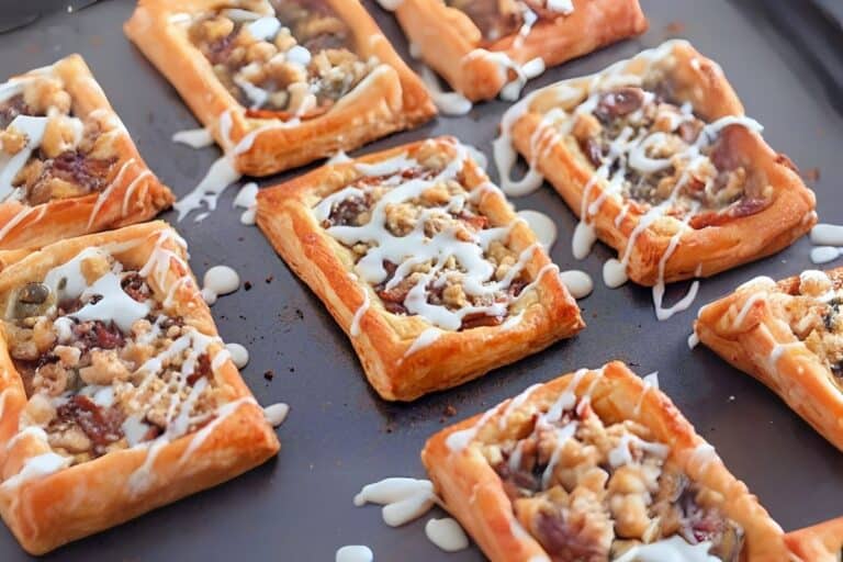 Golden-brown Dutch apple danishes topped with fruit, crumble topping and drizzled with white frosting on a dark baking sheet.