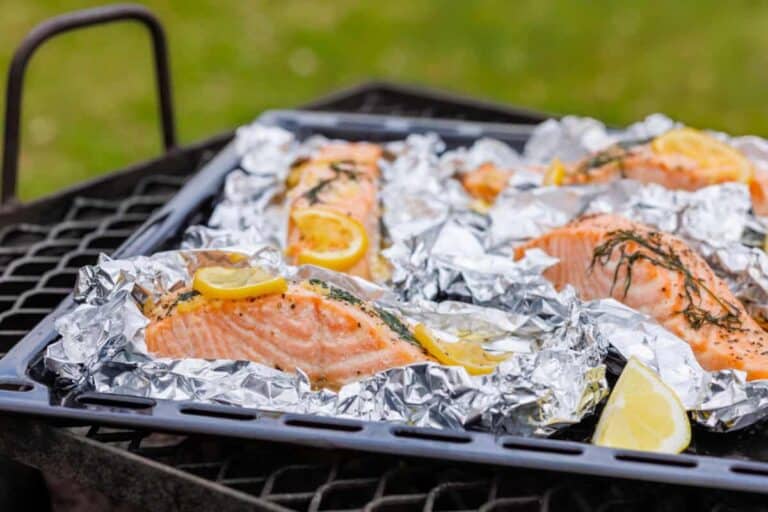 Several salmon fillets with lemon slices and herbs are placed in aluminum foil on a grill tray, ready for cooking outdoors.