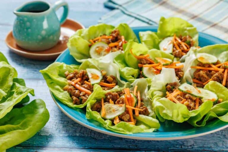 A blue plate with lettuce wraps filled with ground meat, shredded carrots, and cucumber slices. A small pitcher of sauce sits on a saucer nearby.