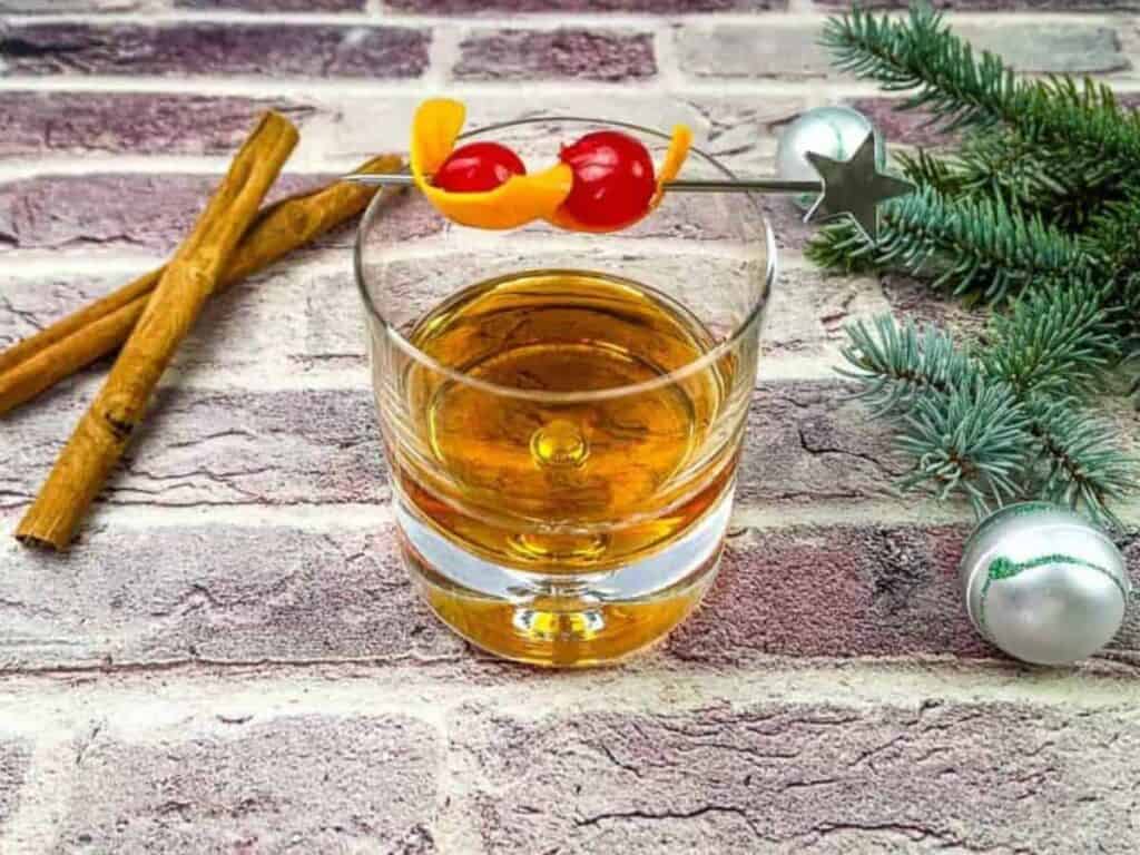 A glass of amber-colored liquor garnished with cherries and orange peel on a skewer, next to cinnamon sticks, fir sprig, and silver ornaments on a brick surface.