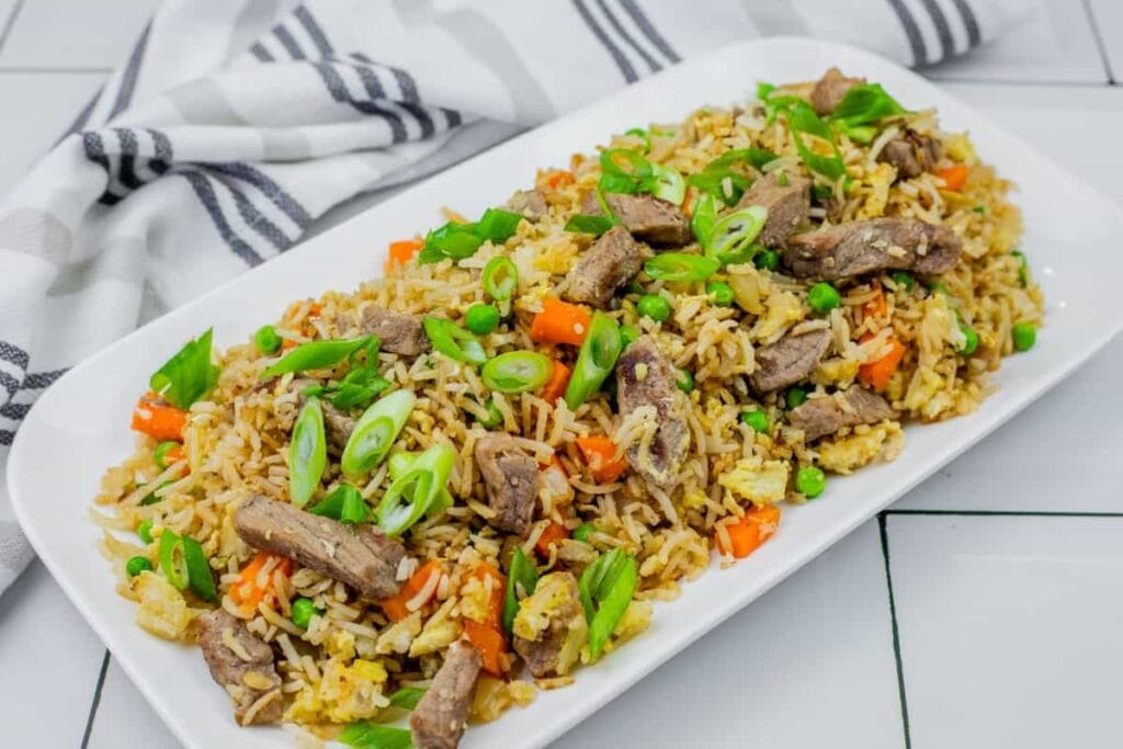 A rectangular white plate with fried rice, sliced beef, peas, carrots, and chopped green onions, placed on a tiled surface with a striped kitchen towel in the background.