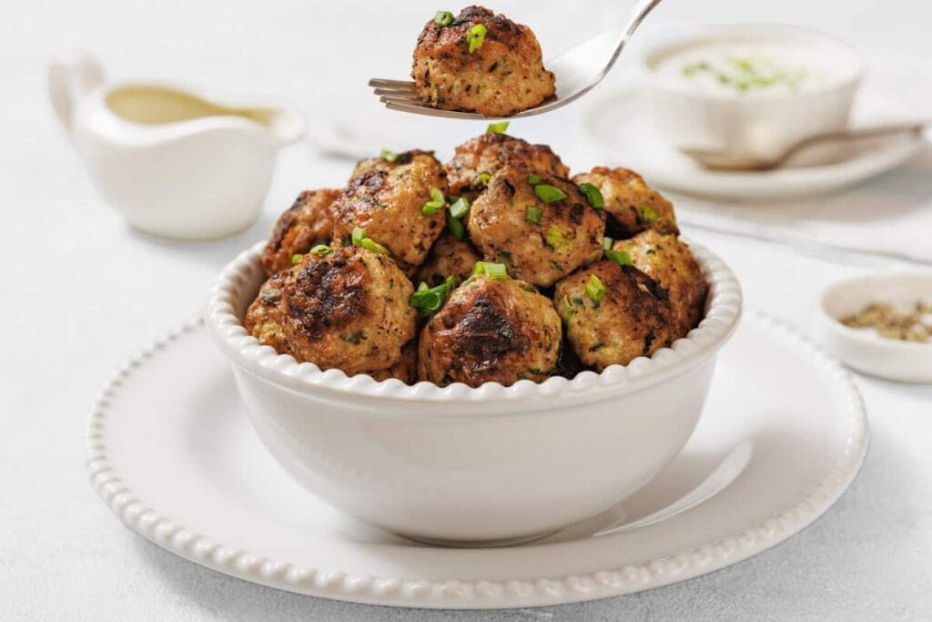 A white bowl filled with cooked meatballs garnished with chopped green herbs, with a fork holding one meatball above the bowl; sauce and seasonings in the background.