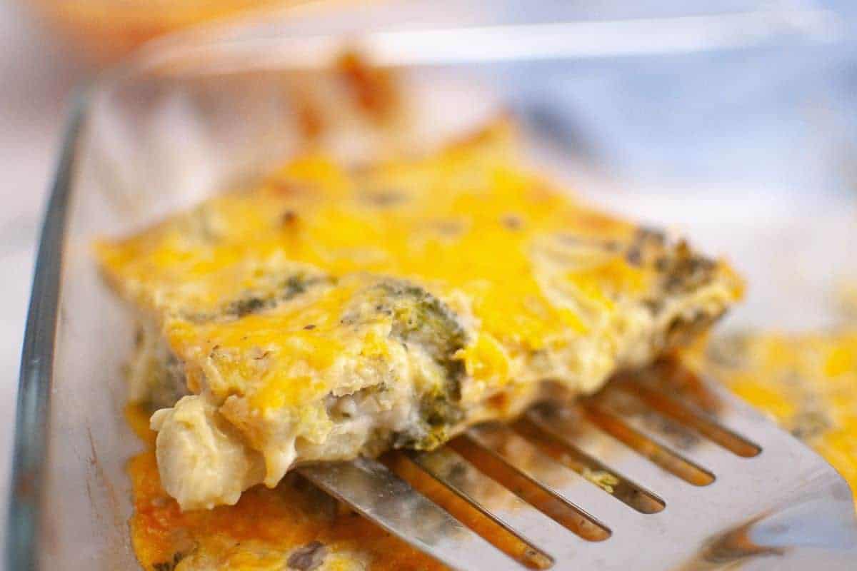 A metal spatula lifts a slice of 3-Ingredient Broccoli Cheese Casserole from a glass baking dish.