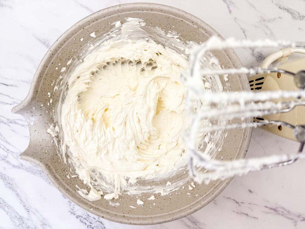 A mixing bowl of whipped cream cheese with an electric hand mixer resting on the side, seen from above on a marble surface.