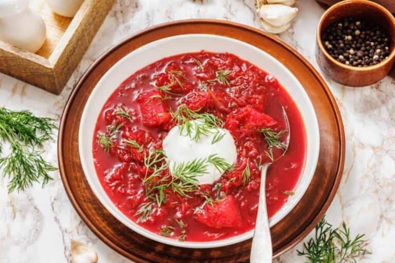 A bowl of red beet soup with chunks of beet, topped with a dollop of sour cream and fresh dill, on a wooden plate with a spoon.