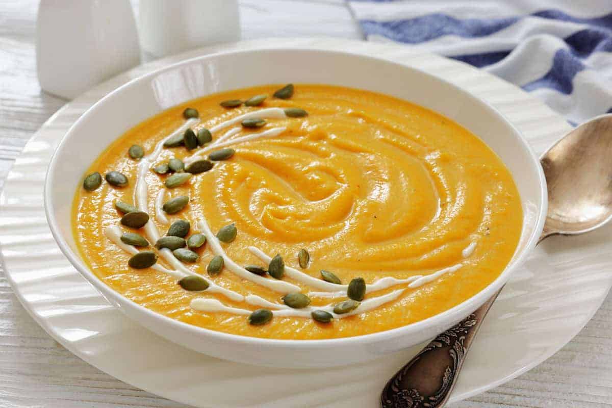 A bowl of creamy pumpkin soup with canned pumpkin garnished with a swirl of cream and sprinkled with pumpkin seeds, placed on a white plate beside a spoon.