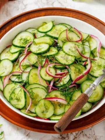 A bowl of cucumber salad with red onions and fresh dill, placed on a wooden plate with a fork, surrounded by ingredients on a marble surface.