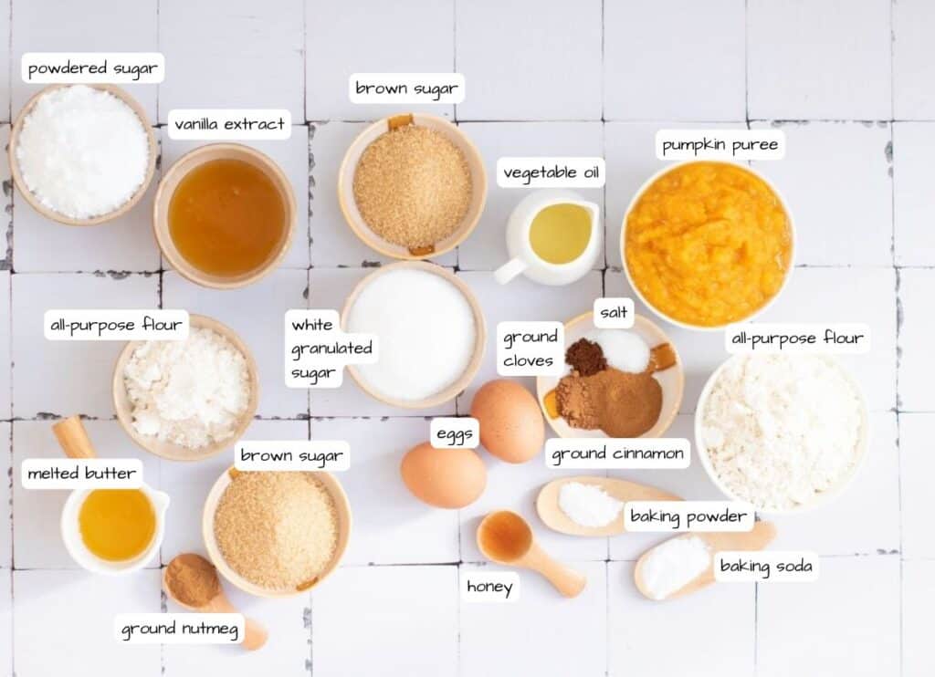 A top view of various labeled baking ingredients in bowls and spoons on a white surface, including flours, sugars, spices, eggs, and pumpkin puree.