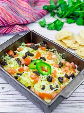 A black baking dish filled with layered dip topped with shredded lettuce, diced tomatoes, sliced black olives, and a jalape&ntilde;o slice. Tortilla chips and fresh cilantro are in the background.