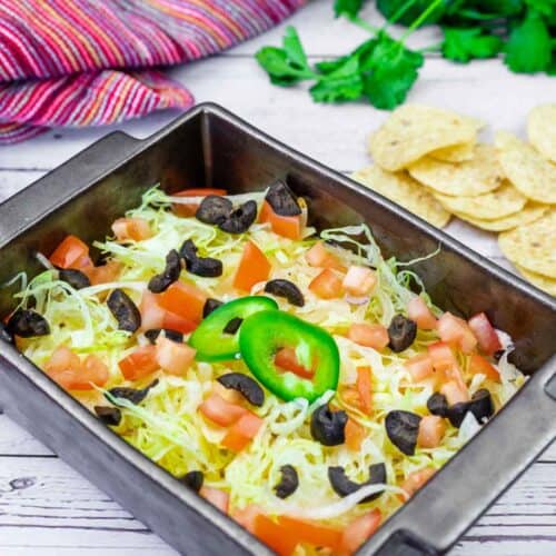 A black baking dish filled with layered dip topped with shredded lettuce, diced tomatoes, sliced black olives, and a jalapeño slice. Tortilla chips and fresh cilantro are in the background.